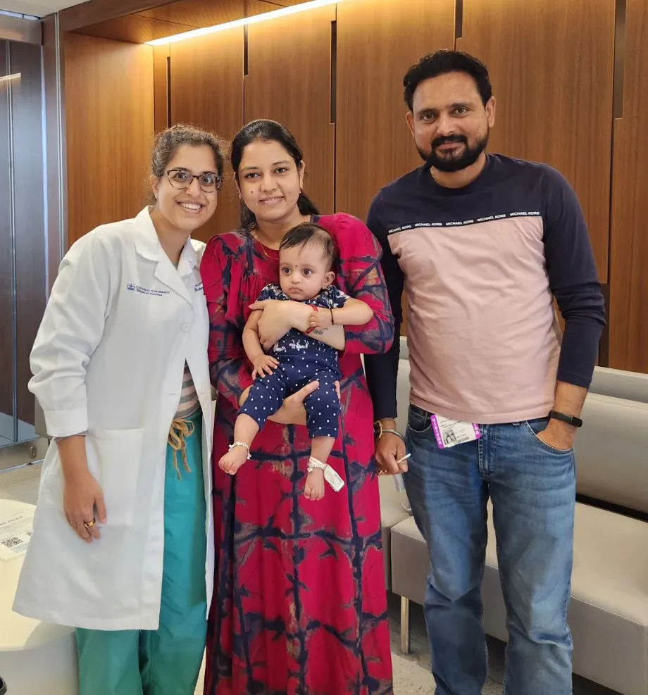 A family of three poses with a female doctor in a hospital setting. The mother, dressed in a vibrant pink dress, holds a baby wearing a blue polka-dotted outfit, while the father stands to the right in casual attire. The doctor, wearing a white lab coat and green scrubs, smiles warmly beside them. The background features wooden paneling and seating.