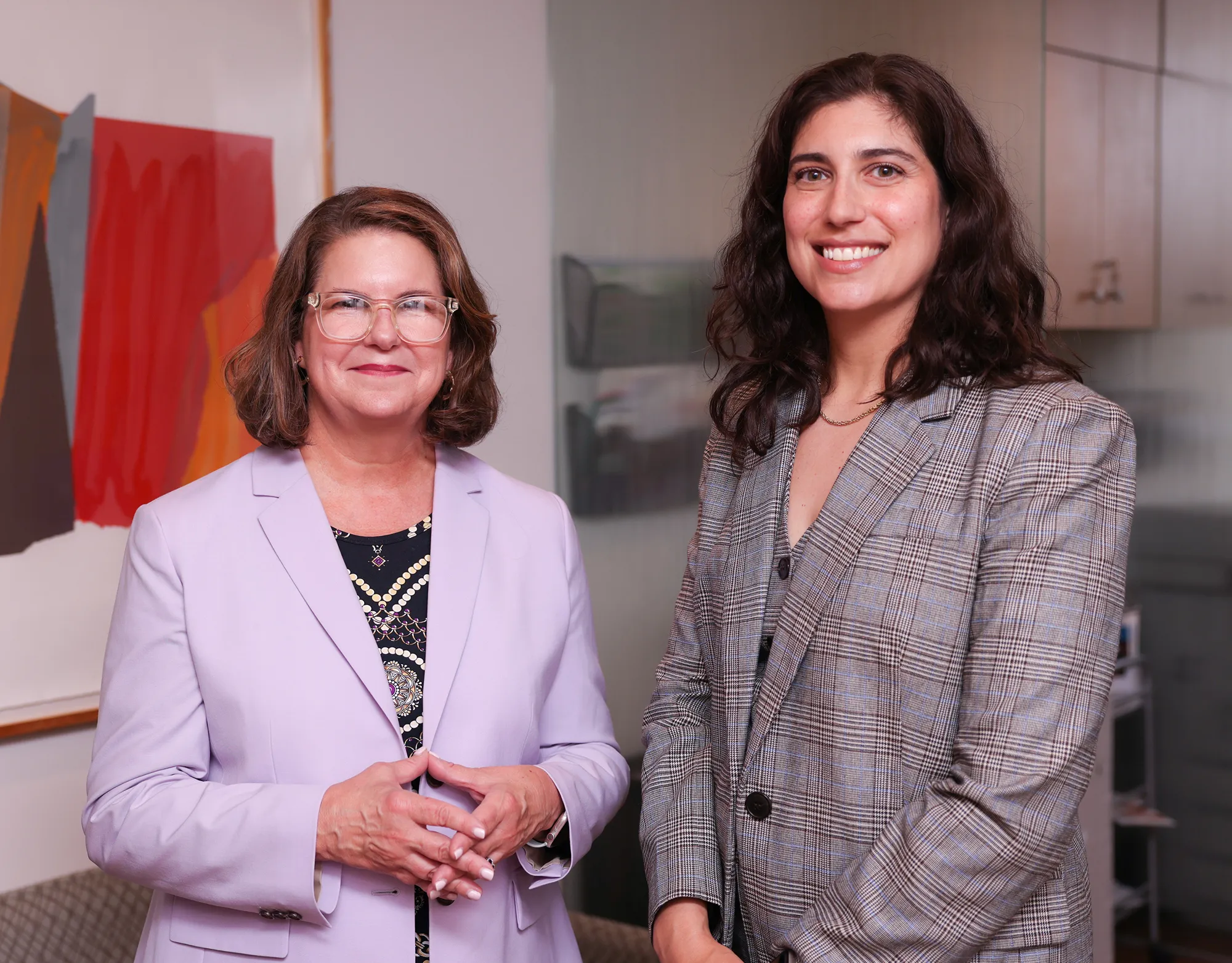 Two women, one in a lavender blazer and the other in a plaid jacket, pose together in an office setting. Both women are smiling, and abstract artwork is visible on the wall behind them.